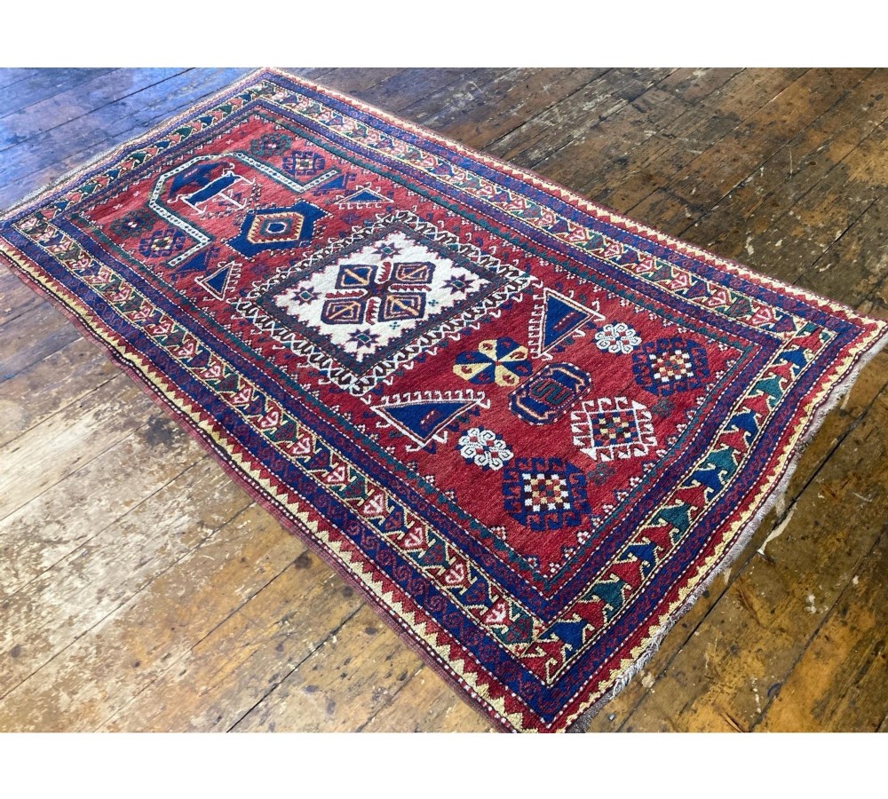 A hand-knotted Caucasian prayer rug featuring a red central field with a pointed mihrab niche, geometric medallions, and multi-band patterned borders in navy blue, red, and cream. View V