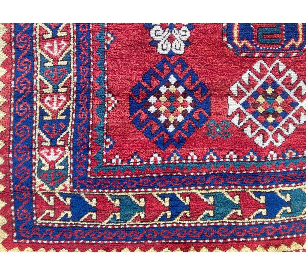 A hand-knotted Caucasian prayer rug featuring a red central field with a pointed mihrab niche, geometric medallions, and multi-band patterned borders in navy blue, red, and cream. View VI