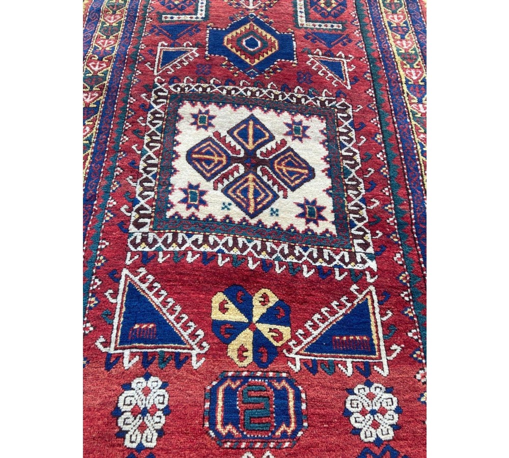 A hand-knotted Caucasian prayer rug featuring a red central field with a pointed mihrab niche, geometric medallions, and multi-band patterned borders in navy blue, red, and cream. View VII