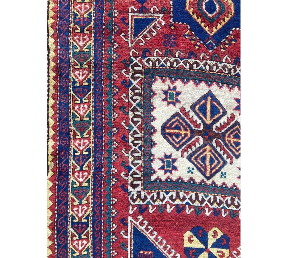 A hand-knotted Caucasian prayer rug featuring a red central field with a pointed mihrab niche, geometric medallions, and multi-band patterned borders in navy blue, red, and cream. View XII