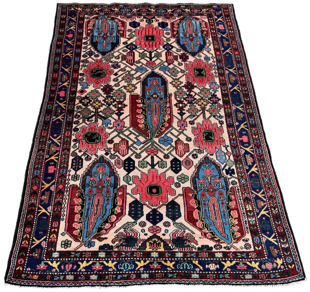 An antique Persian Bakhtiar wool rug featuring intricate geometric floral patterns and bird motifs in blue, pink, and red set against a cream background with a dark blue decorative border. View II