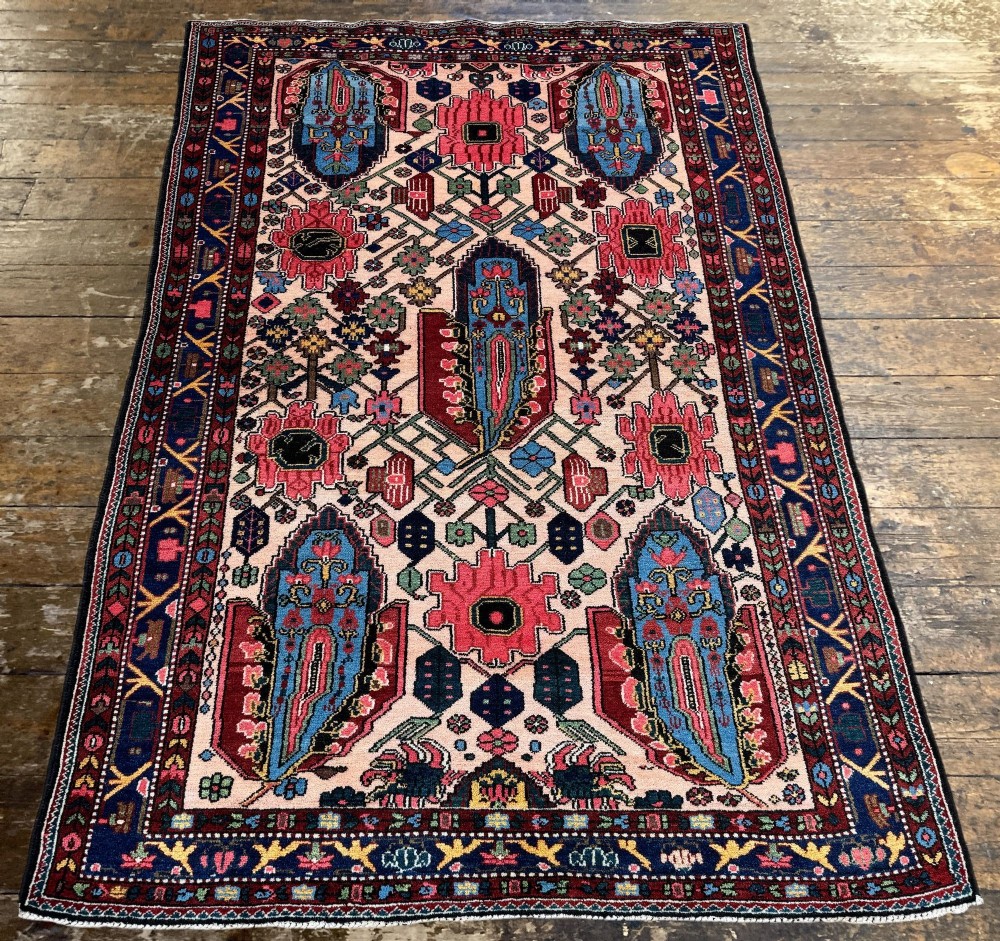 An antique Persian Bakhtiar wool rug featuring intricate geometric floral patterns and bird motifs in blue, pink, and red set against a cream background with a dark blue decorative border. View IV