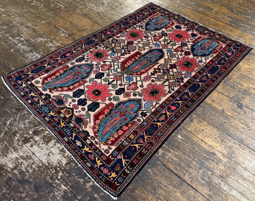 An antique Persian Bakhtiar wool rug featuring intricate geometric floral patterns and bird motifs in blue, pink, and red set against a cream background with a dark blue decorative border. View V