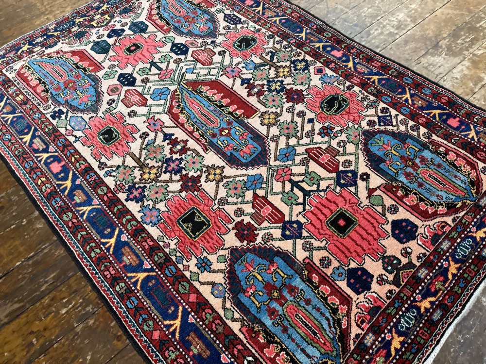 An antique Persian Bakhtiar wool rug featuring intricate geometric floral patterns and bird motifs in blue, pink, and red set against a cream background with a dark blue decorative border. View VI