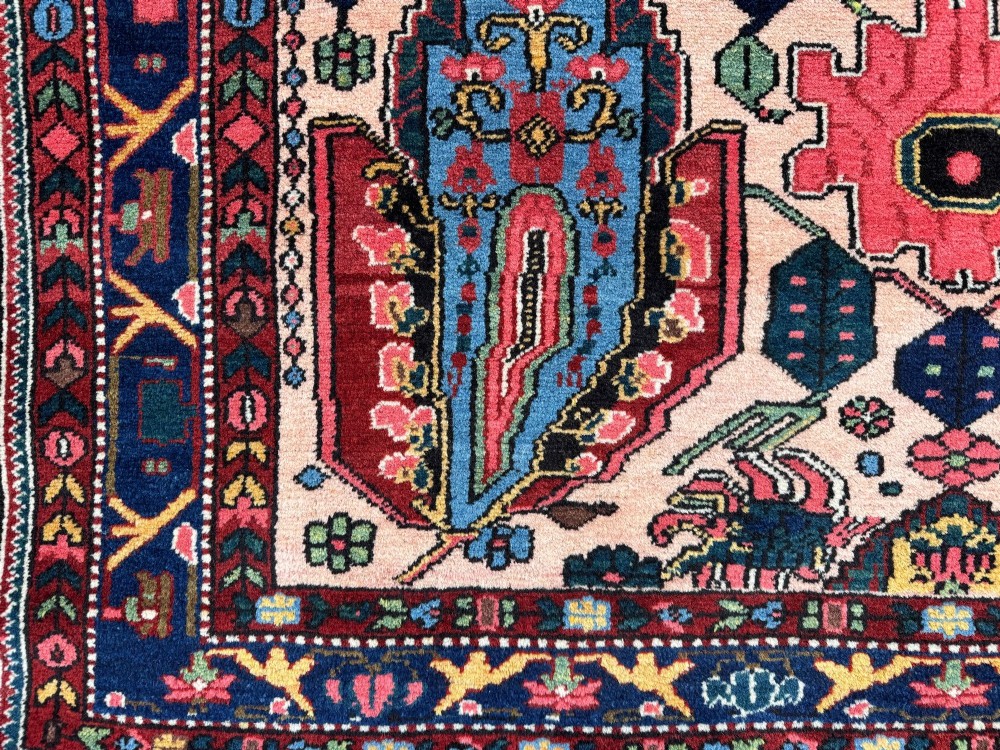 An antique Persian Bakhtiar wool rug featuring intricate geometric floral patterns and bird motifs in blue, pink, and red set against a cream background with a dark blue decorative border. View VII