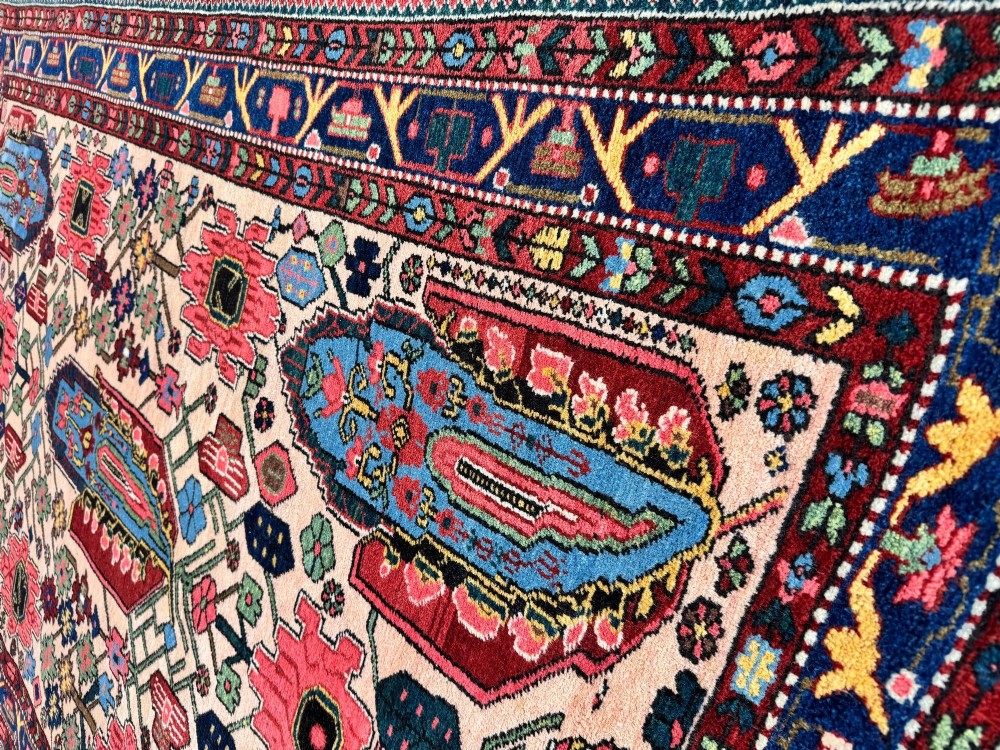 An antique Persian Bakhtiar wool rug featuring intricate geometric floral patterns and bird motifs in blue, pink, and red set against a cream background with a dark blue decorative border. View IX