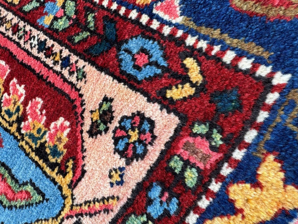 An antique Persian Bakhtiar wool rug featuring intricate geometric floral patterns and bird motifs in blue, pink, and red set against a cream background with a dark blue decorative border. View X