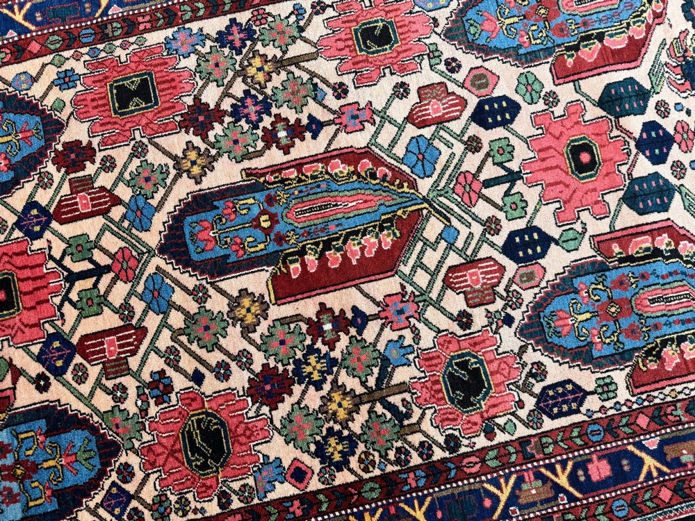 An antique Persian Bakhtiar wool rug featuring intricate geometric floral patterns and bird motifs in blue, pink, and red set against a cream background with a dark blue decorative border. View XI