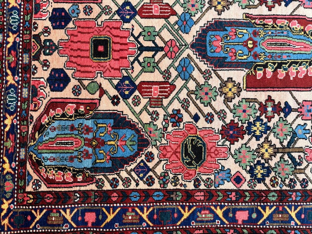 An antique Persian Bakhtiar wool rug featuring intricate geometric floral patterns and bird motifs in blue, pink, and red set against a cream background with a dark blue decorative border. View XII