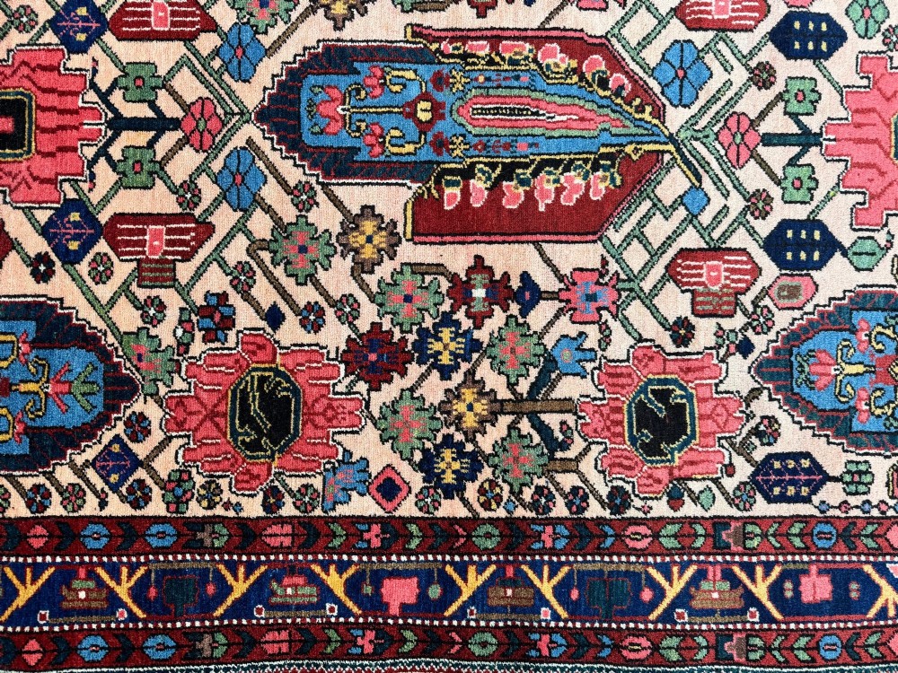 An antique Persian Bakhtiar wool rug featuring intricate geometric floral patterns and bird motifs in blue, pink, and red set against a cream background with a dark blue decorative border. View XIII