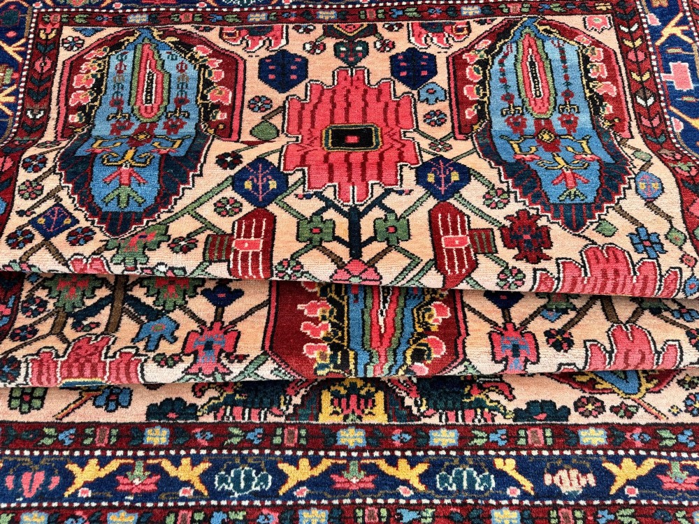 An antique Persian Bakhtiar wool rug featuring intricate geometric floral patterns and bird motifs in blue, pink, and red set against a cream background with a dark blue decorative border. View XIV