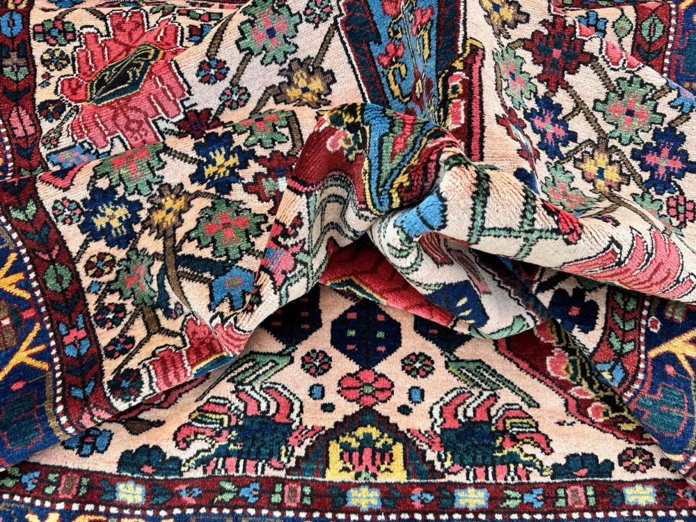 An antique Persian Bakhtiar wool rug featuring intricate geometric floral patterns and bird motifs in blue, pink, and red set against a cream background with a dark blue decorative border. View XV
