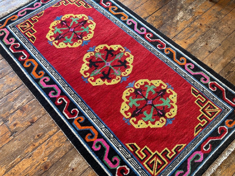 A hand-knotted rectangular Tibetan wool rug featuring a bright red centre field with three yellow and green central medallion motifs framed by a black cloud-scroll border. View IV