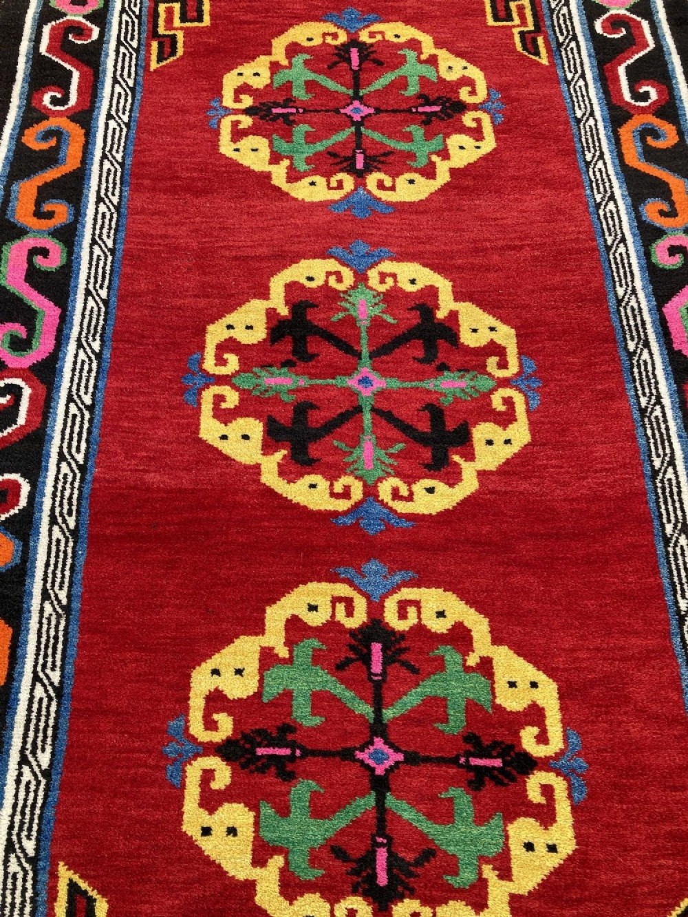 A hand-knotted rectangular Tibetan wool rug featuring a bright red centre field with three yellow and green central medallion motifs framed by a black cloud-scroll border. View VI