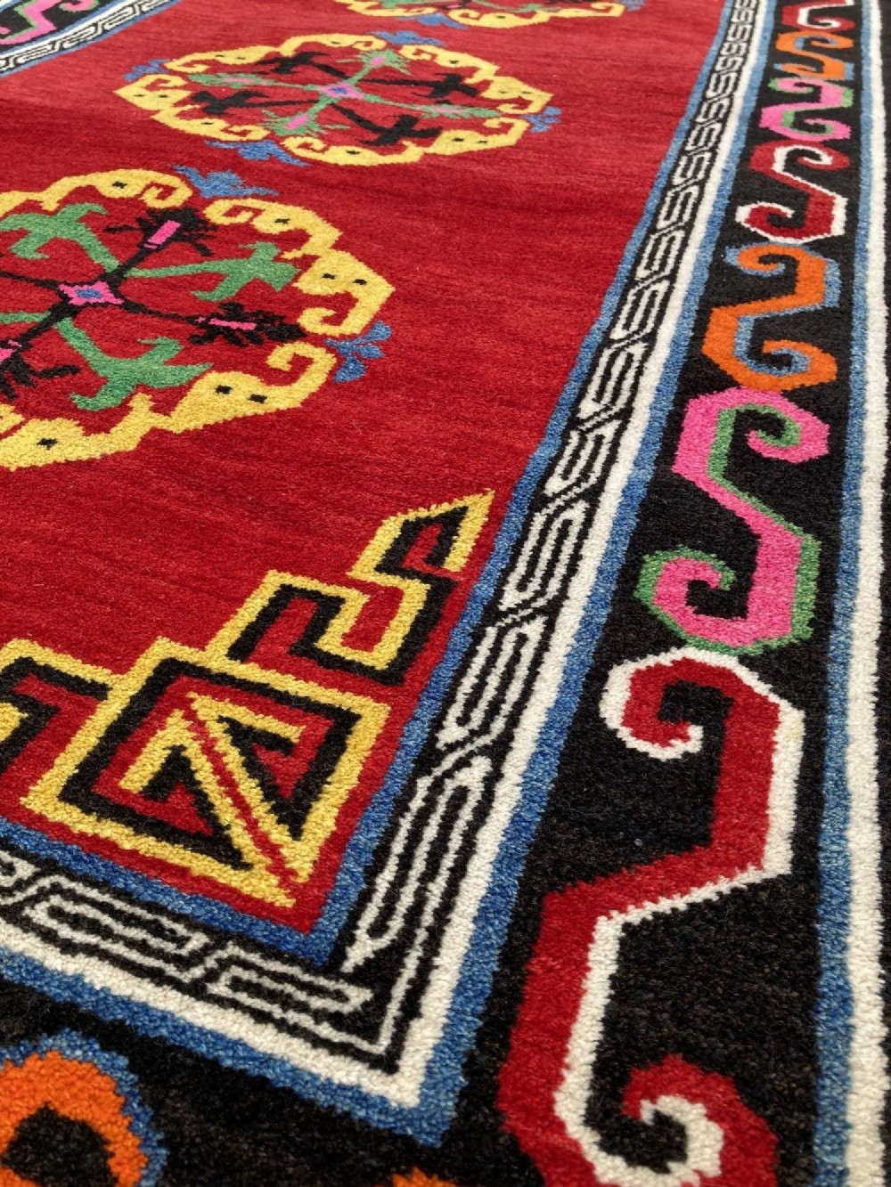 A hand-knotted rectangular Tibetan wool rug featuring a bright red centre field with three yellow and green central medallion motifs framed by a black cloud-scroll border. View VII