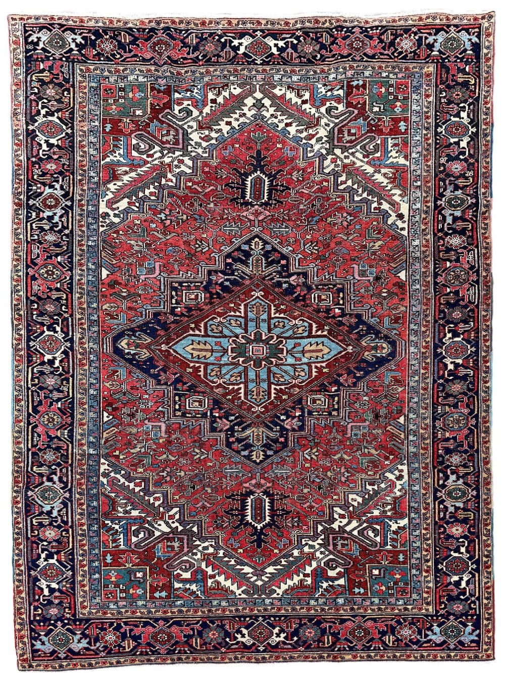 A large hand-knotted antique Heriz wool carpet featuring a prominent central geometric medallion on a deep red field framed by dark navy and cream borders.
