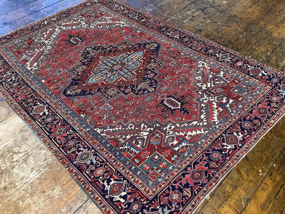 A large hand-knotted antique Heriz wool carpet featuring a prominent central geometric medallion on a deep red field framed by dark navy and cream borders. View IV
