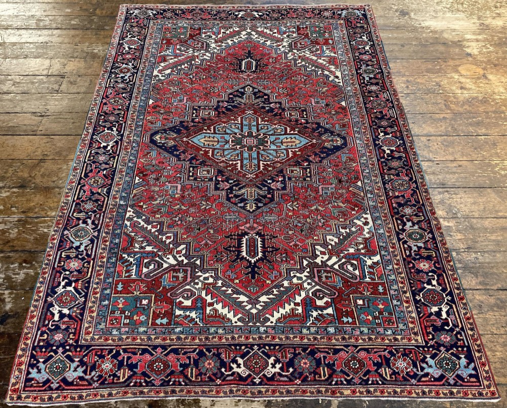 A large hand-knotted antique Heriz wool carpet featuring a prominent central geometric medallion on a deep red field framed by dark navy and cream borders. View V