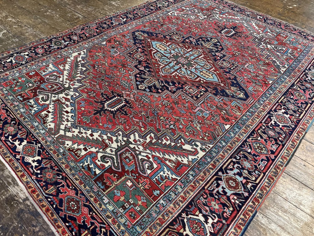 A large hand-knotted antique Heriz wool carpet featuring a prominent central geometric medallion on a deep red field framed by dark navy and cream borders. View VI