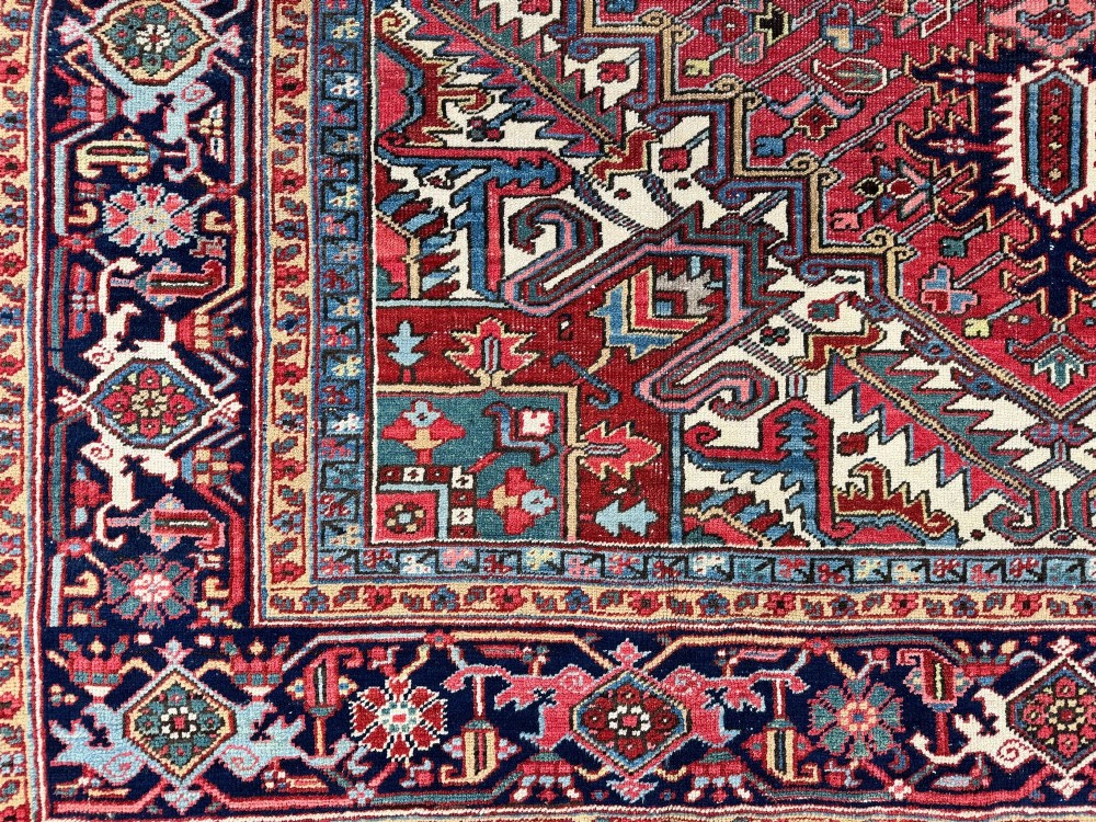 A large hand-knotted antique Heriz wool carpet featuring a prominent central geometric medallion on a deep red field framed by dark navy and cream borders. View VII