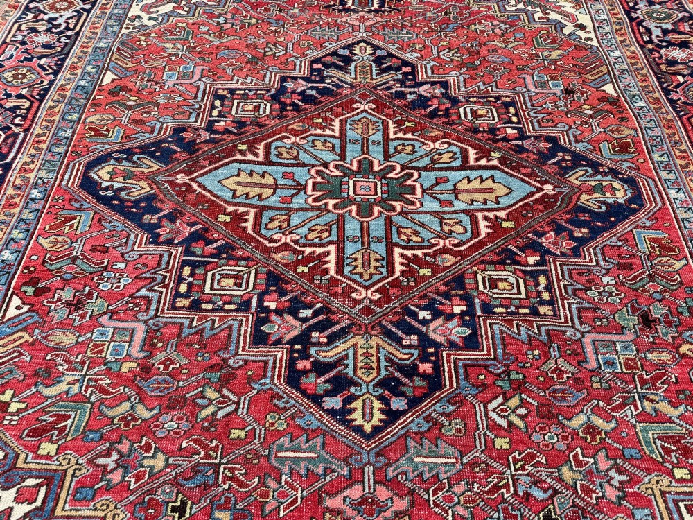A large hand-knotted antique Heriz wool carpet featuring a prominent central geometric medallion on a deep red field framed by dark navy and cream borders. View VIII