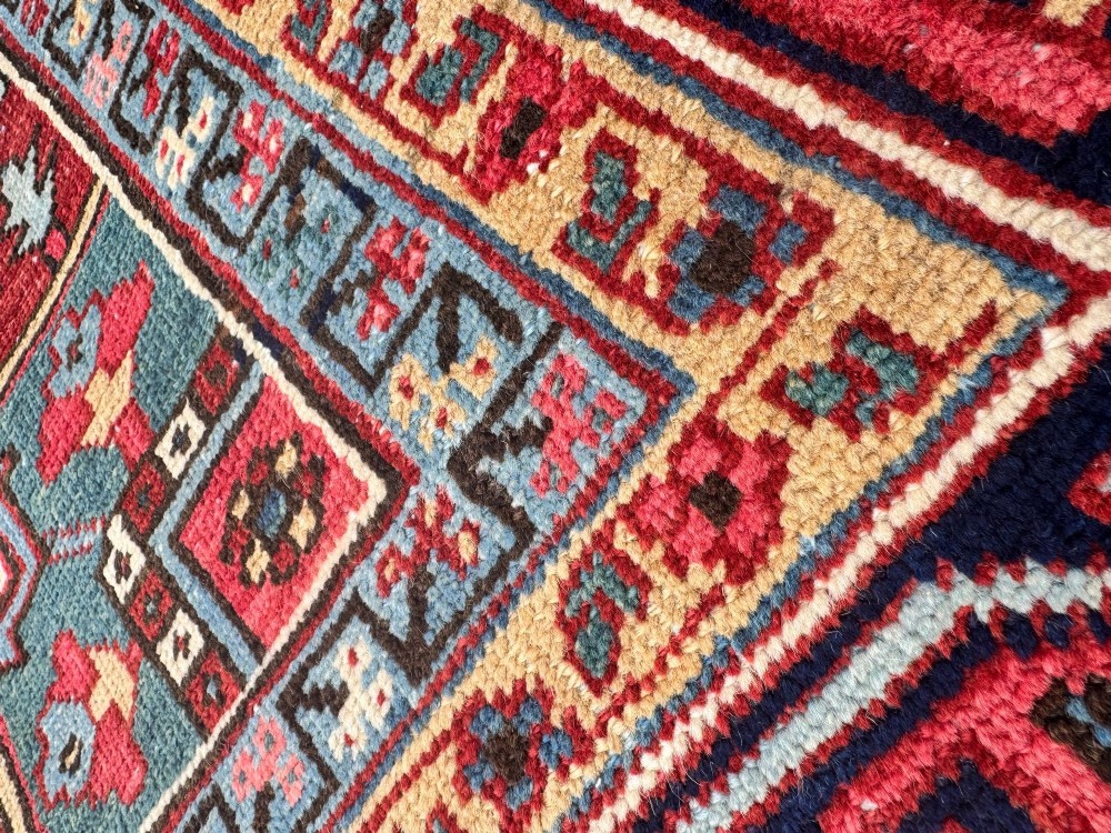 A large hand-knotted antique Heriz wool carpet featuring a prominent central geometric medallion on a deep red field framed by dark navy and cream borders. View X