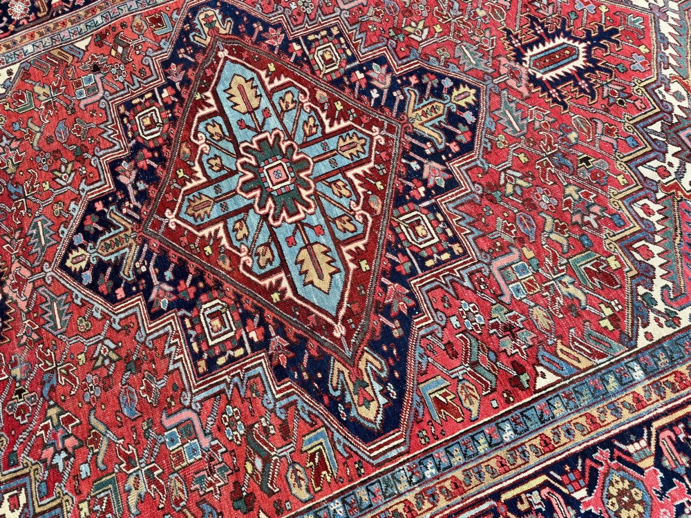 A large hand-knotted antique Heriz wool carpet featuring a prominent central geometric medallion on a deep red field framed by dark navy and cream borders. View XI