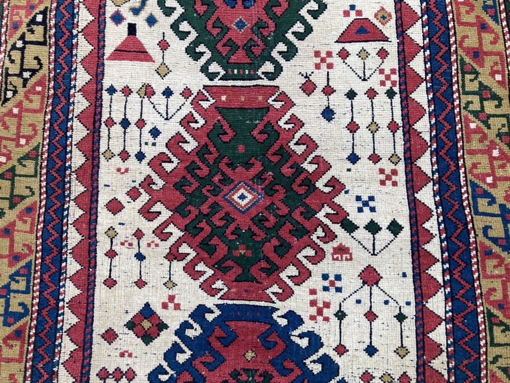 An antique hand-knotted Caucasian Kazak wool rug featuring three stacked geometric medallions on an ivory ground with multi-coloured geometric borders. View VI