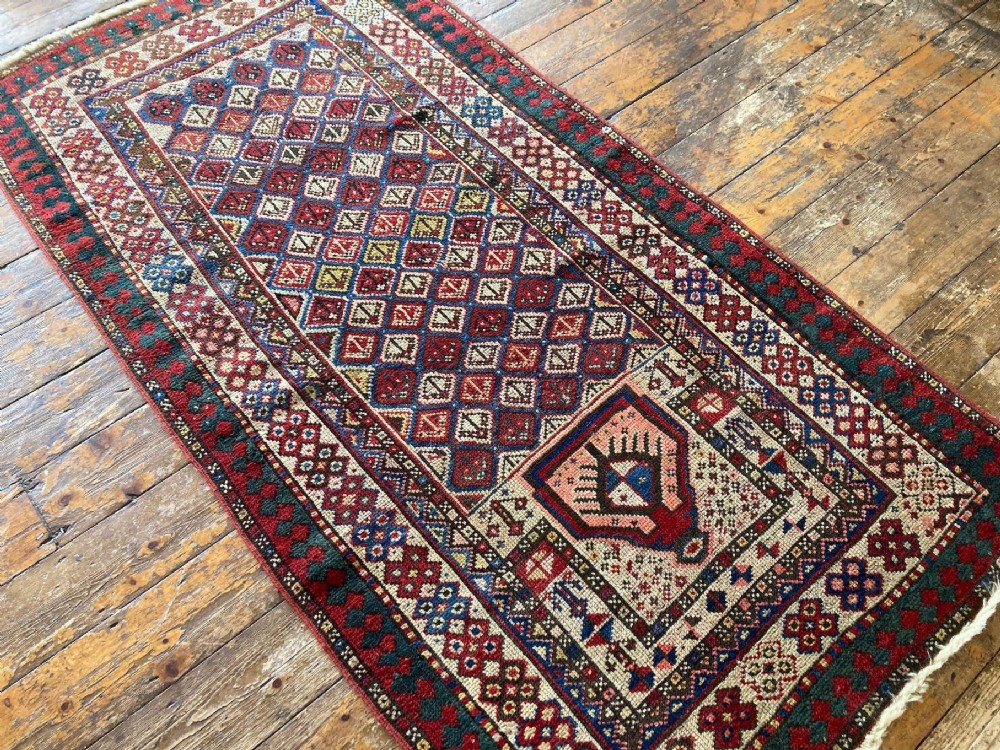 A hand-knotted antique Caucasian Gendje prayer rug featuring a deep red central field with a diamond lattice pattern, topped with a geometric mihrab motif and framed by multi-coloured decorative borders. View IV