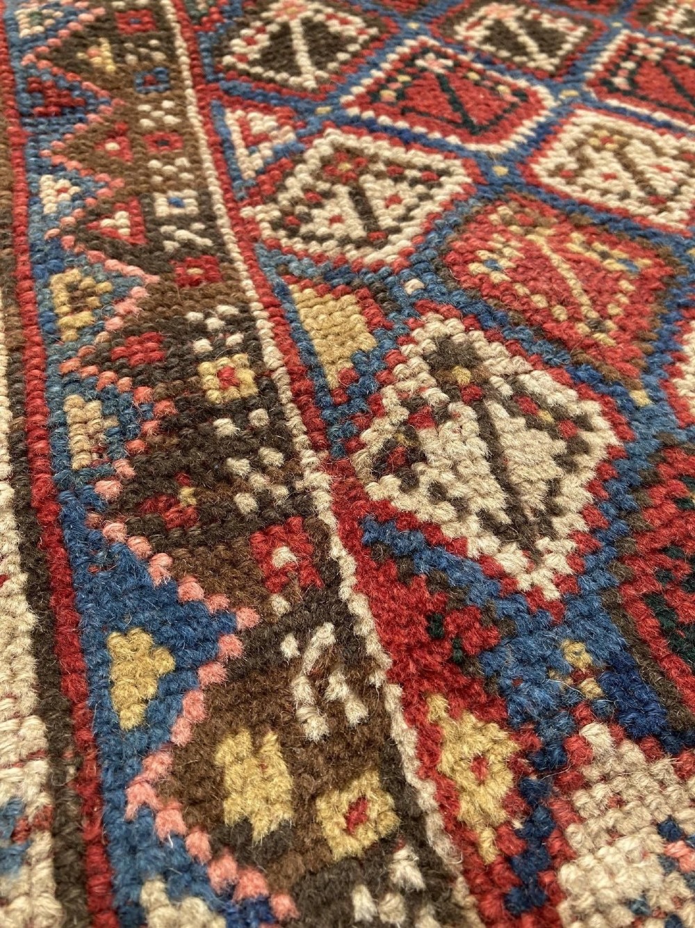 A hand-knotted antique Caucasian Gendje prayer rug featuring a deep red central field with a diamond lattice pattern, topped with a geometric mihrab motif and framed by multi-coloured decorative borders. View IX