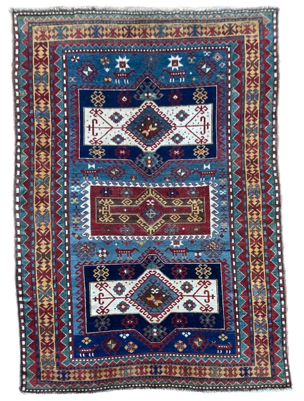 An antique Caucasian Kazak wool rug featuring three central geometric medallions surrounded by stylised animal figures and multi-tiered patterned borders in blue, red, cream, and yellow hues.