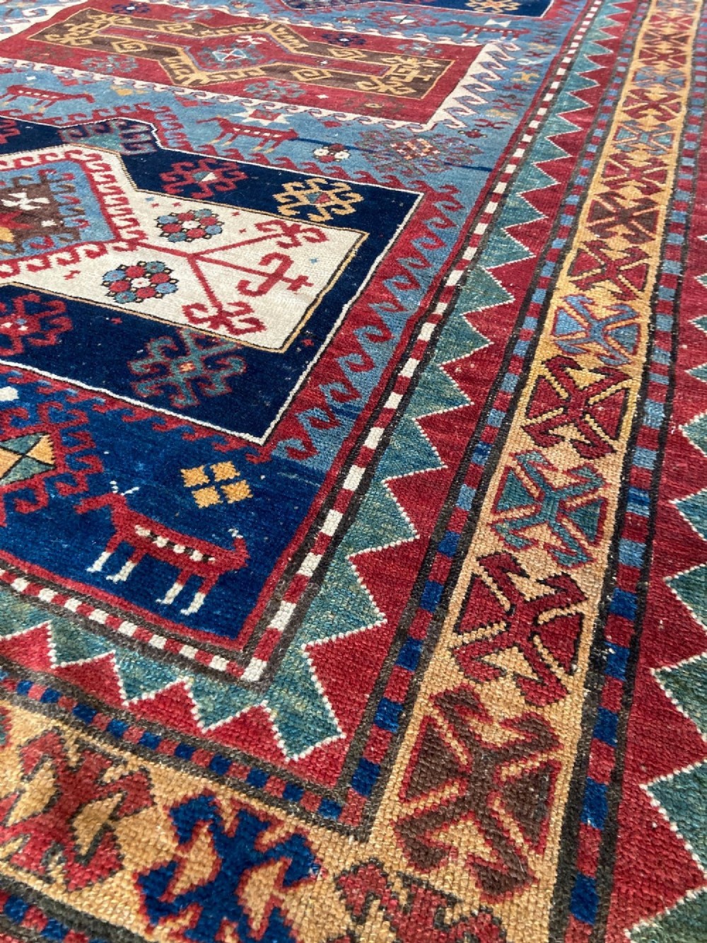 An antique Caucasian Kazak wool rug featuring three central geometric medallions surrounded by stylised animal figures and multi-tiered patterned borders in blue, red, cream, and yellow hues. View VIII