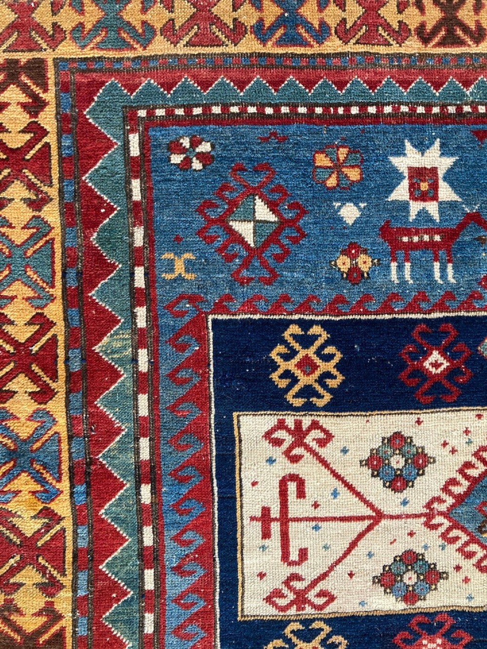An antique Caucasian Kazak wool rug featuring three central geometric medallions surrounded by stylised animal figures and multi-tiered patterned borders in blue, red, cream, and yellow hues. View X