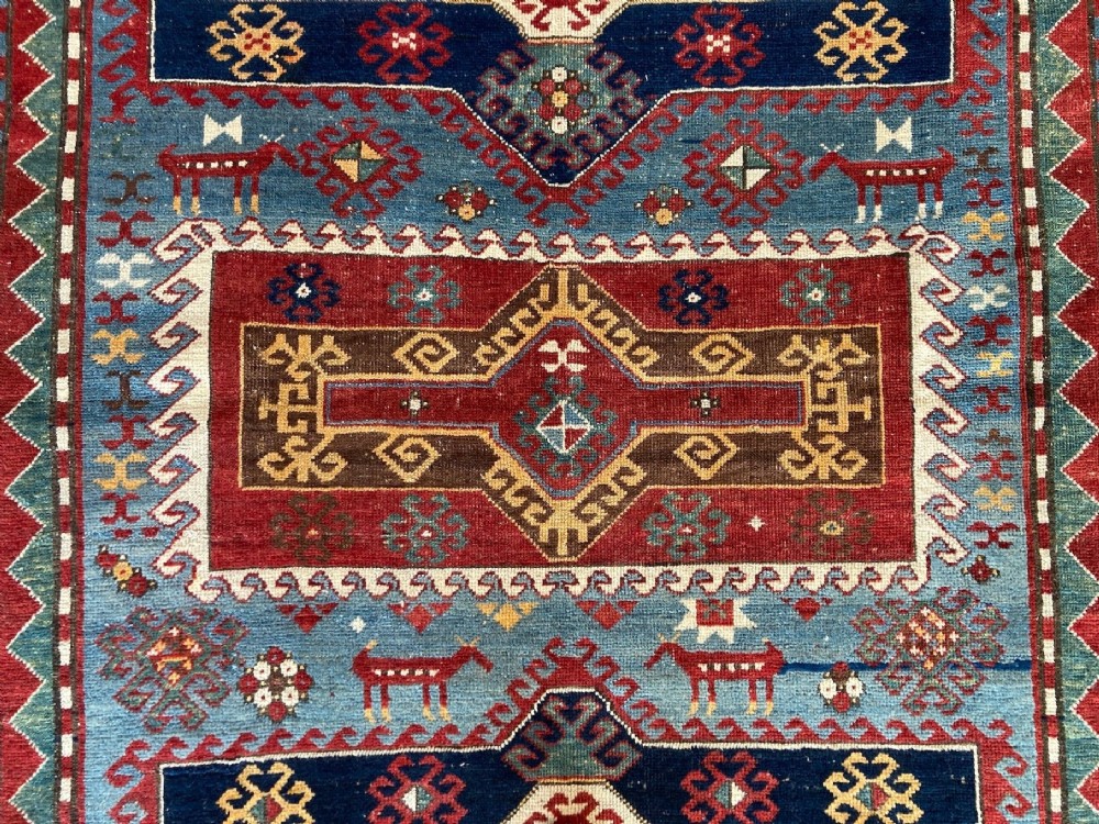 An antique Caucasian Kazak wool rug featuring three central geometric medallions surrounded by stylised animal figures and multi-tiered patterned borders in blue, red, cream, and yellow hues. View XIII