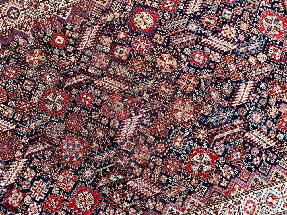 An antique Shekarlu tribal wool rug featuring dense dark navy and red geometric motifs enclosed by a patterned ivory border. View X