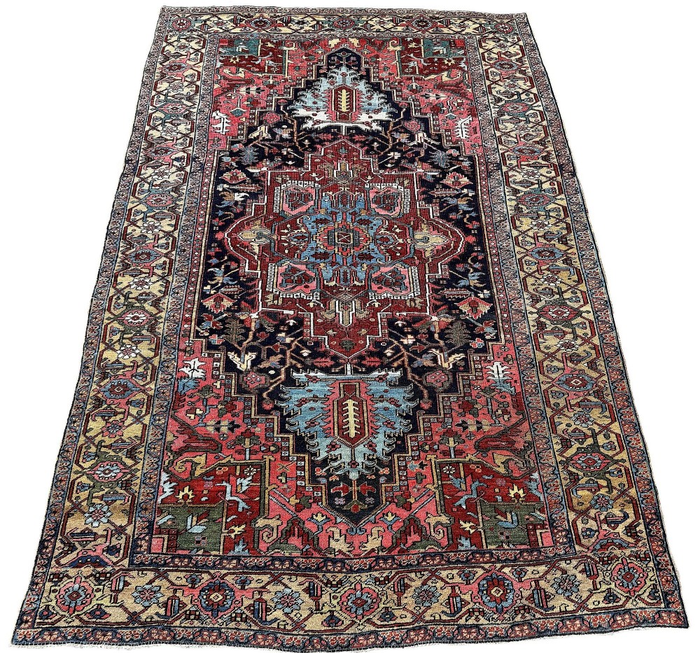 An antique Persian Heriz wool carpet featuring a large central geometric medallion in navy blue and light blue, set against a terracotta red field with broad angular floral borders. View II