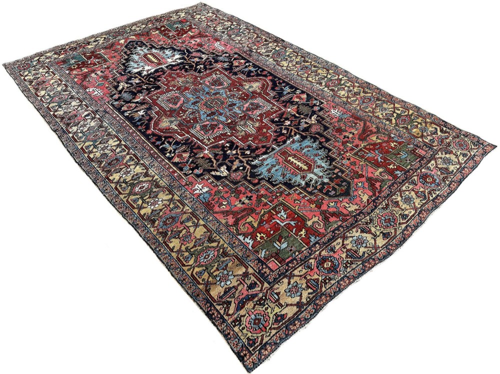 An antique Persian Heriz wool carpet featuring a large central geometric medallion in navy blue and light blue, set against a terracotta red field with broad angular floral borders. View III
