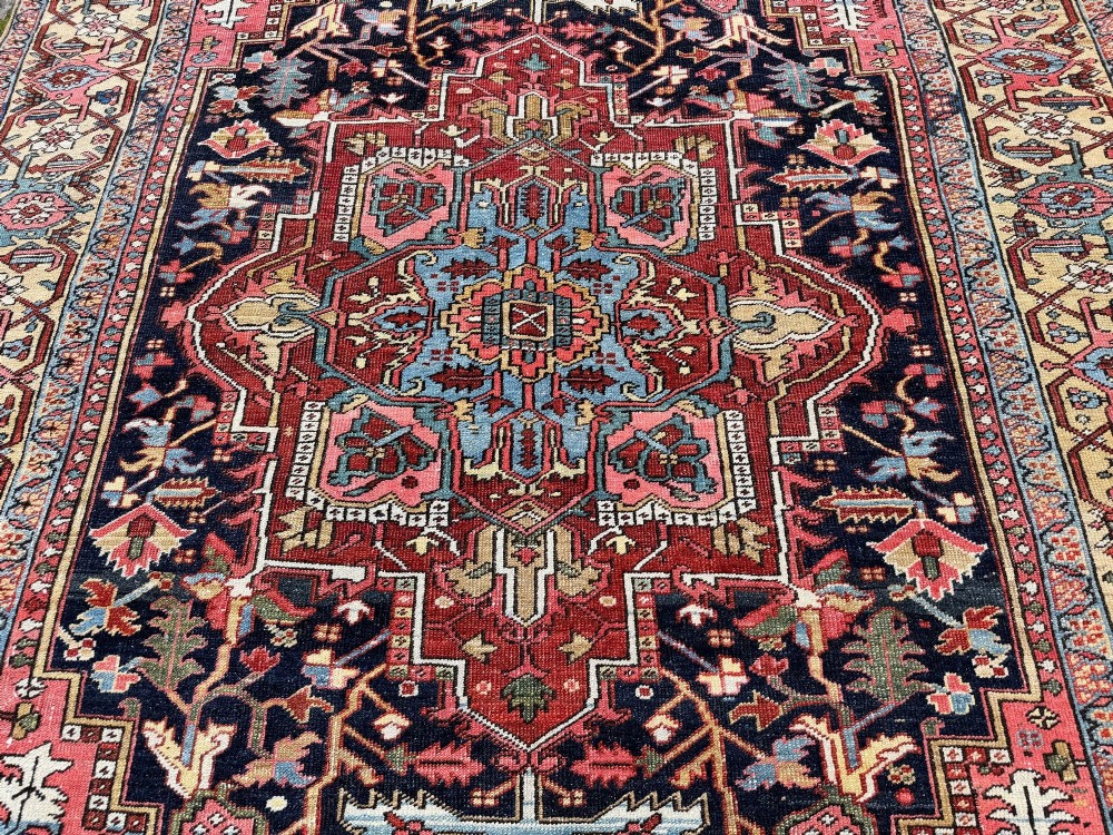 An antique Persian Heriz wool carpet featuring a large central geometric medallion in navy blue and light blue, set against a terracotta red field with broad angular floral borders. View VIII