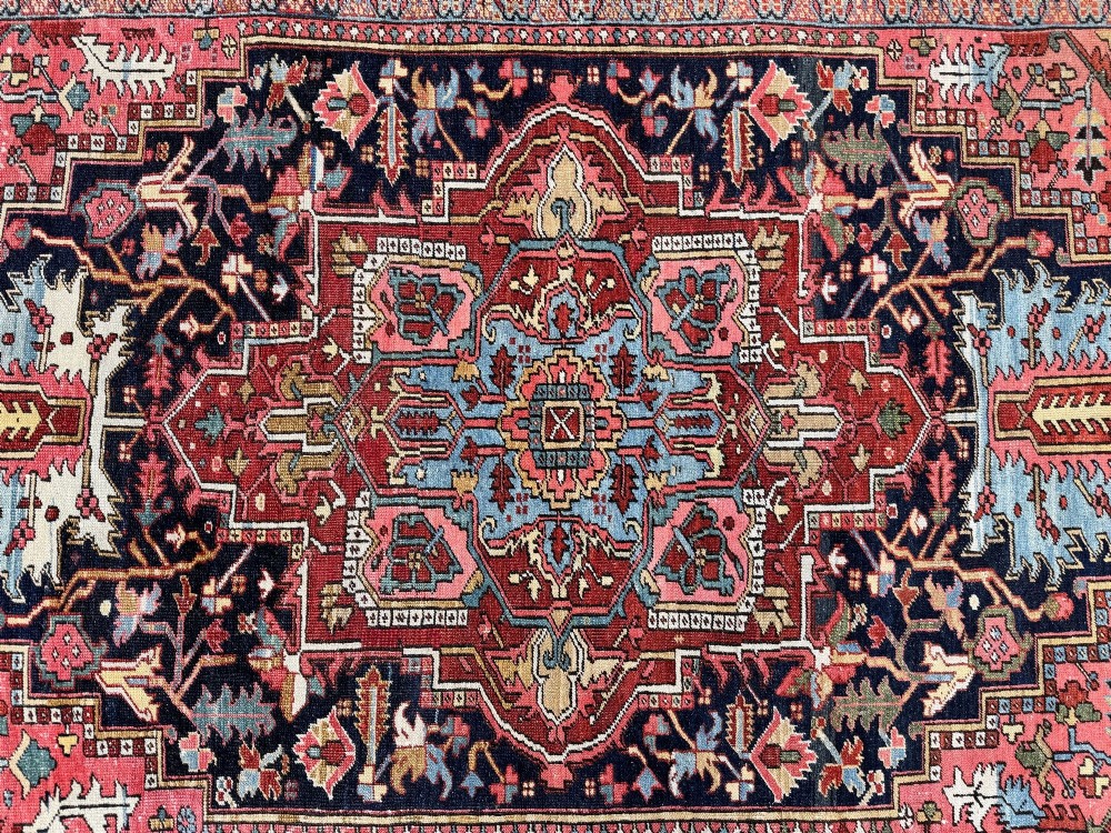 An antique Persian Heriz wool carpet featuring a large central geometric medallion in navy blue and light blue, set against a terracotta red field with broad angular floral borders. View XIV