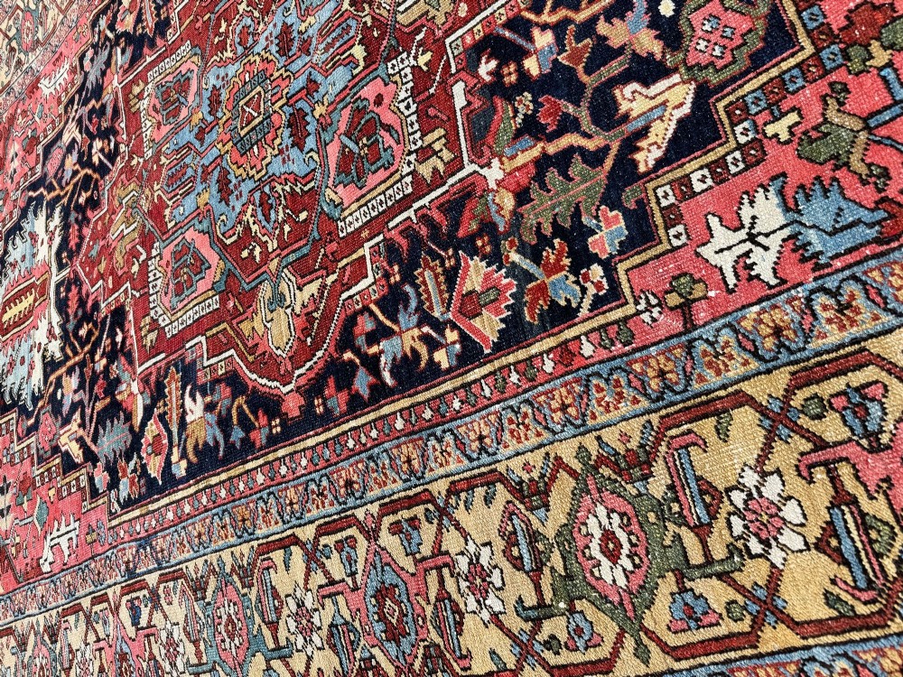An antique Persian Heriz wool carpet featuring a large central geometric medallion in navy blue and light blue, set against a terracotta red field with broad angular floral borders. View XV