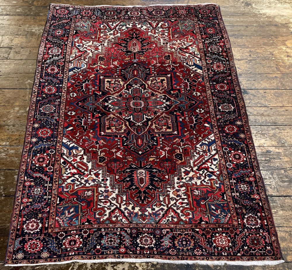 An antique hand-knotted Persian Heriz carpet featuring a prominent geometric central medallion on a rust red field with cream corner spandrels and a navy blue border. View III