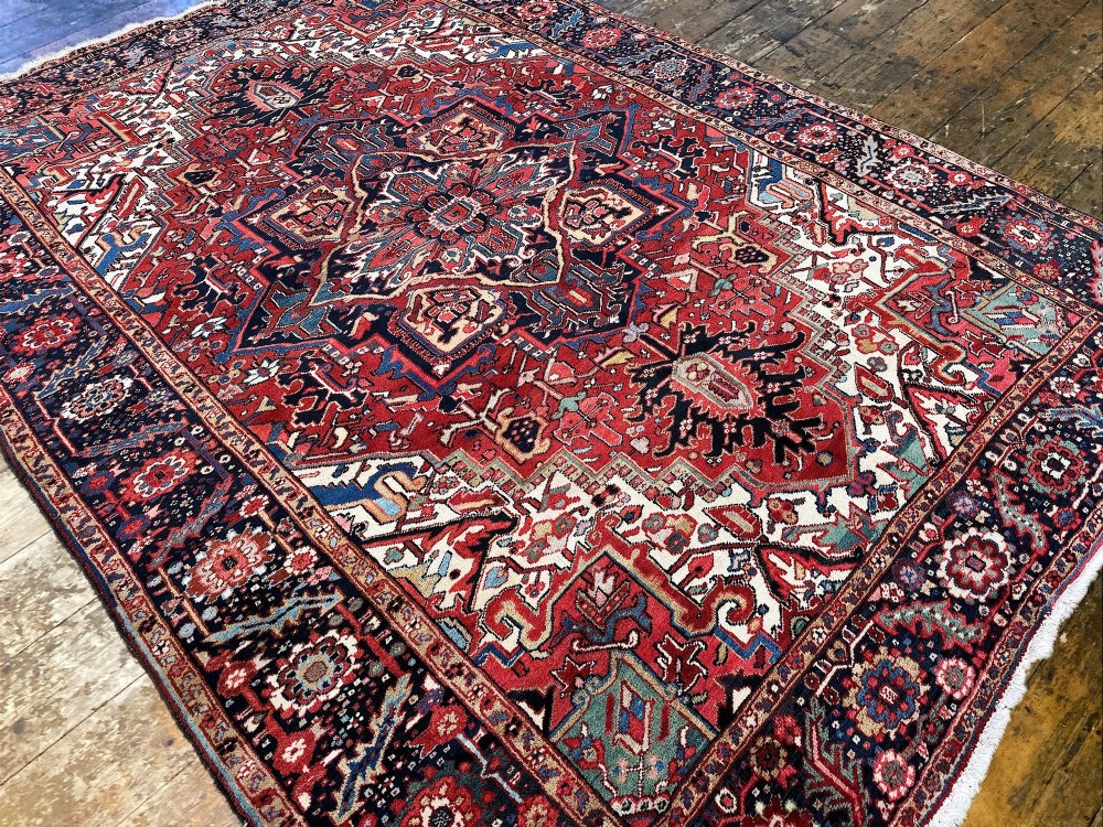 An antique hand-knotted Persian Heriz carpet featuring a prominent geometric central medallion on a rust red field with cream corner spandrels and a navy blue border. View V