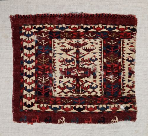 fig 1. A hand-woven wool Turkmen Teke mafrash textile fragment featuring geometric motifs in deep red, blue, and cream, mounted on a light beige backing.