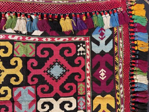 fig 1. A square antique Central Asian Lakai embroidered textile featuring bold geometric scroll motifs in yellow, magenta, white, and blue surrounded by multi-coloured tassel fringe.