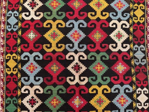 fig 1. A square Uzbek Lakai tribal textile bag front featuring geometric multicolored hook patterns surrounded by a fringed deep red border.