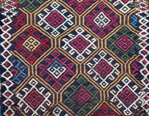 fig 1. A square flatweave textile bag featuring geometric diamond motifs in red, yellow, green, magenta, and white wool against a dark background with a braided cord.