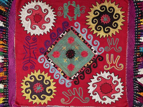 fig 1. A square red silk embroidered textile featuring central diamond and sunburst motifs bordered by dense multi-coloured thread tassels.