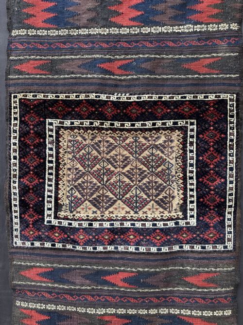 fig 1. An antique handwoven Baluch Sofreh flatweave textile featuring geometric chevron bands in dark red and navy framing a central panel with tree motifs.