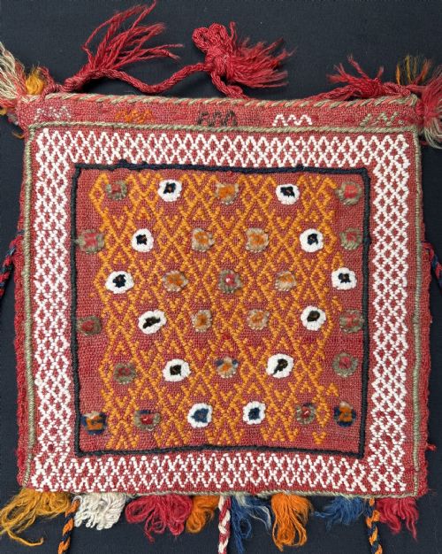 fig 1. A small square woven wool Qashqai vanity bag featuring a red geometric patterned centre, braided borders, and multi-coloured hanging tassels.