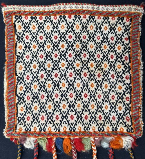 fig 1. A square woven Persian tribal vanity bag featuring a black and red geometric diamond pattern on a cream background, finished with woven borders and long braided tassels ending in colourful fringes.